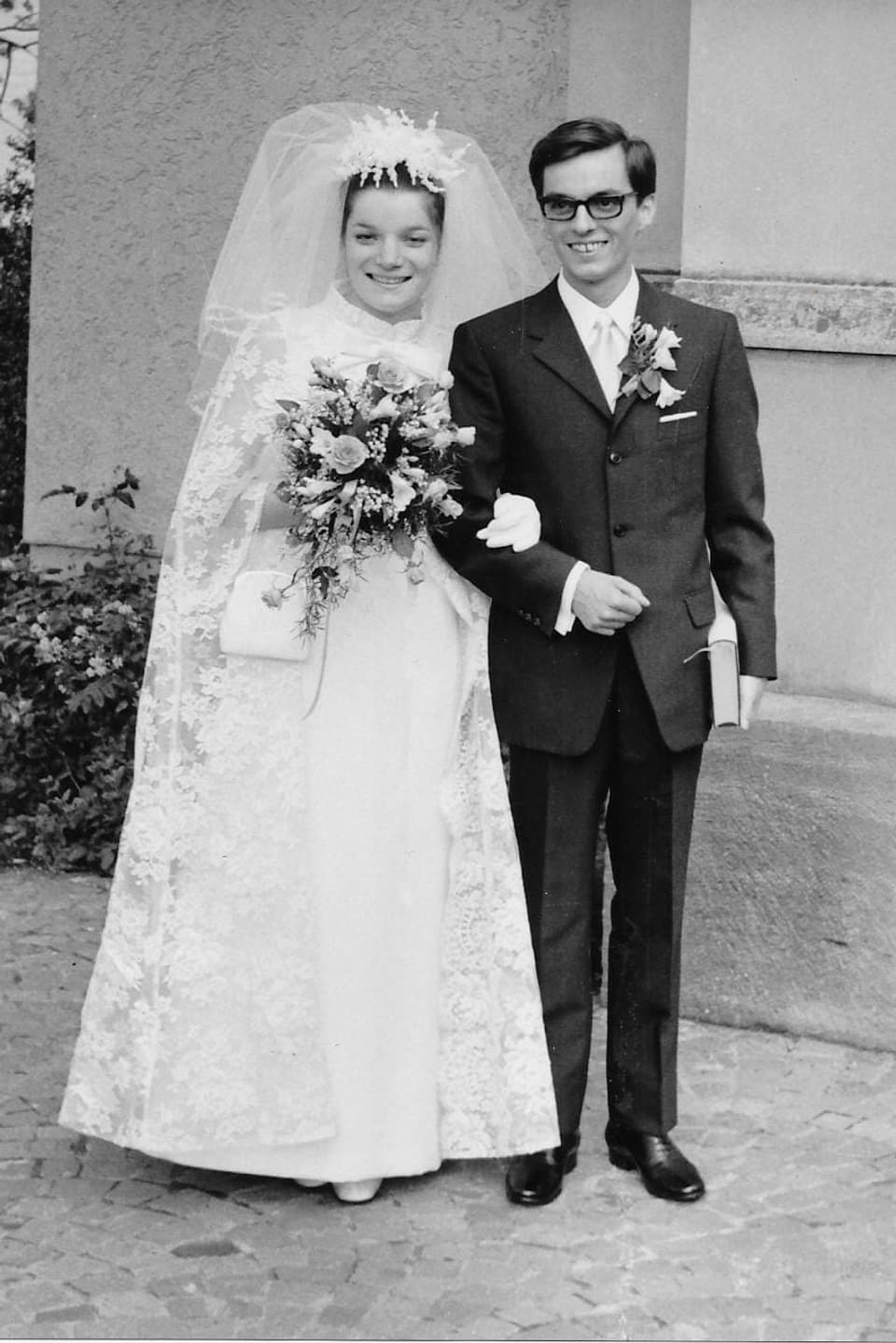 Ein Schwarzweiss-Foto eines Brautpaars. Sie im weissen Kleid mit Spitzen-Umhang und Schleier, er im dunklen Anzug mit Blumen im Knopfloch.