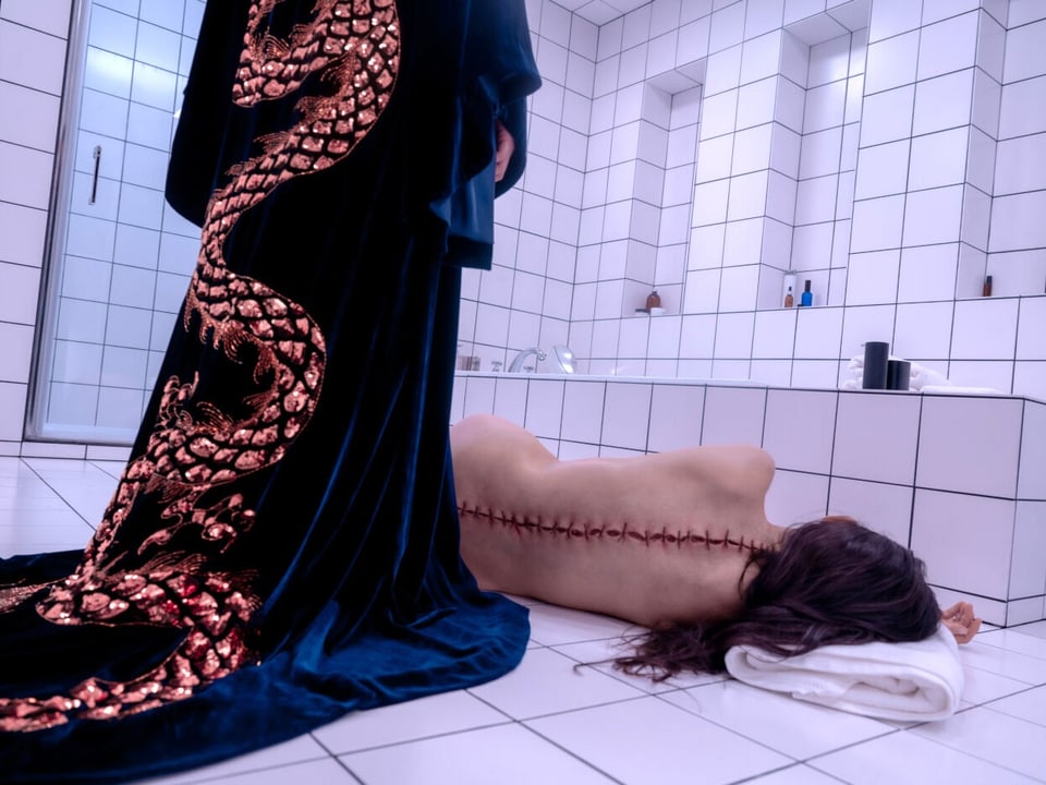Uma mulher está nua com as costas costuradas no chão de um banheiro de azulejos brancos.