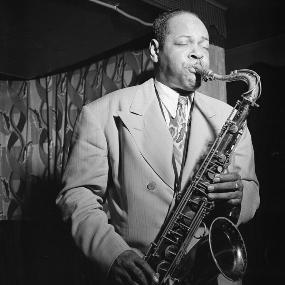 Ein Mann in Anzug und Krawatte auf einer Bühne, Saxophon spielend.