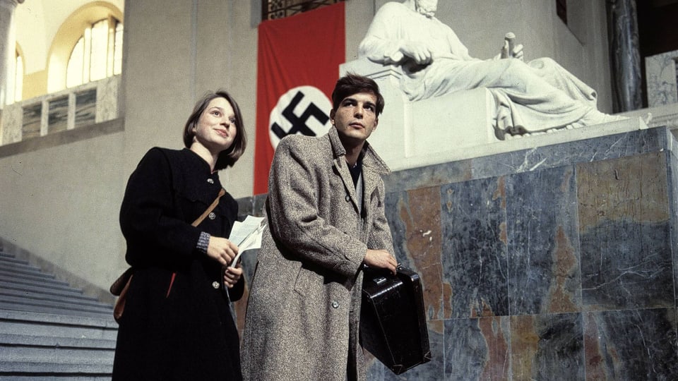 Mann und Frau stehen vor einer Statue und einem Hakenkreuz-Banner in einem historischen Gebäude.