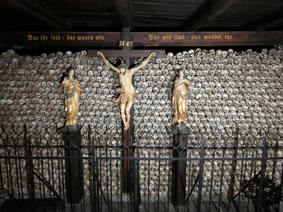 Figur von Jesus am Kreuz, dahinter Wand mit Totenschädeln