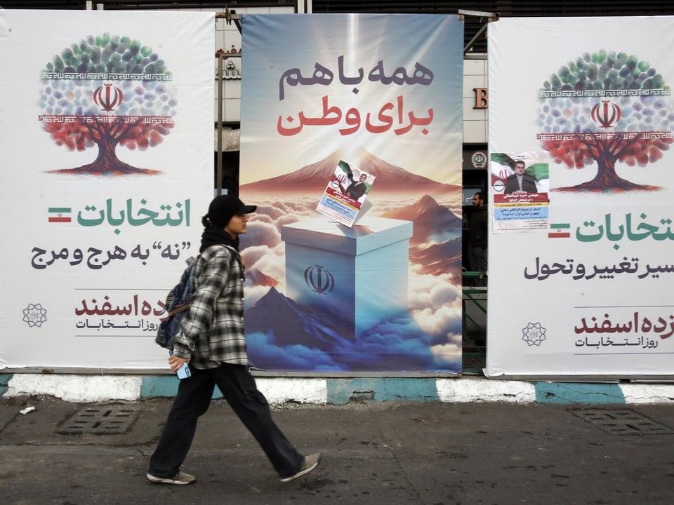 Eine Frau mit Baseballcap läuft vor drei Wahlplakaten vorbei mit persischer Schrift.