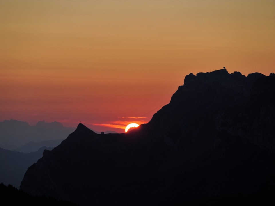 Aufgehende rote Sonnenscheibe, noch halb verdeckt von einer dunklen Bergflanke im Vordergrund.