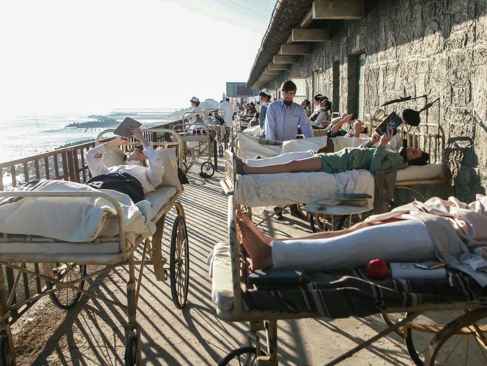 Ein paar Menschen liegen in Krankenbetten auf einer Terrasse, die das Meer überblickt.