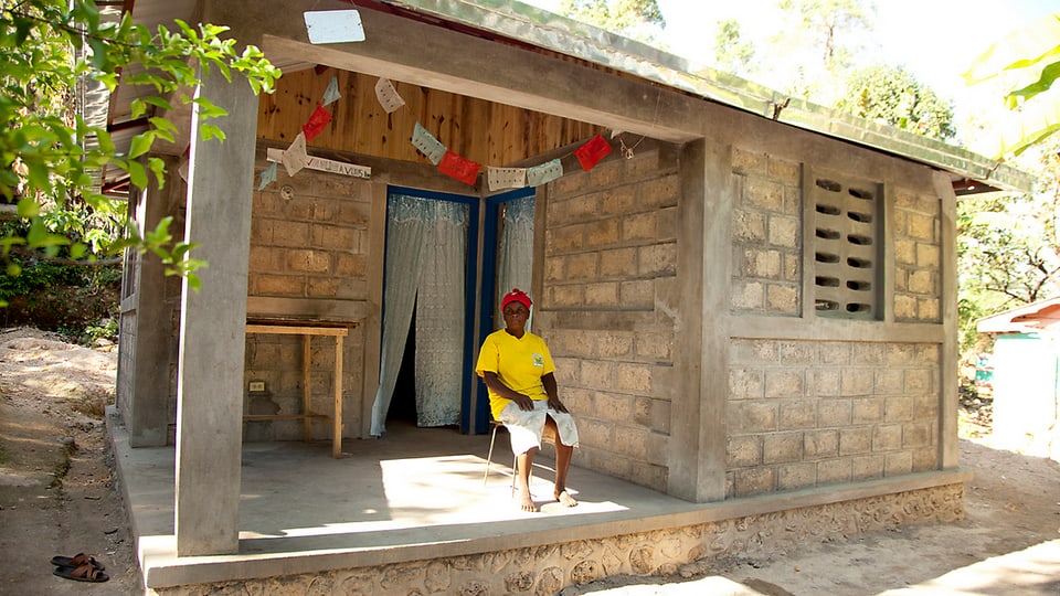 Ein kleines, gemauertes und betoniertes Haus, davor sitzt eine Haitianerin in gelbem T-Shirt, sie lächelt.