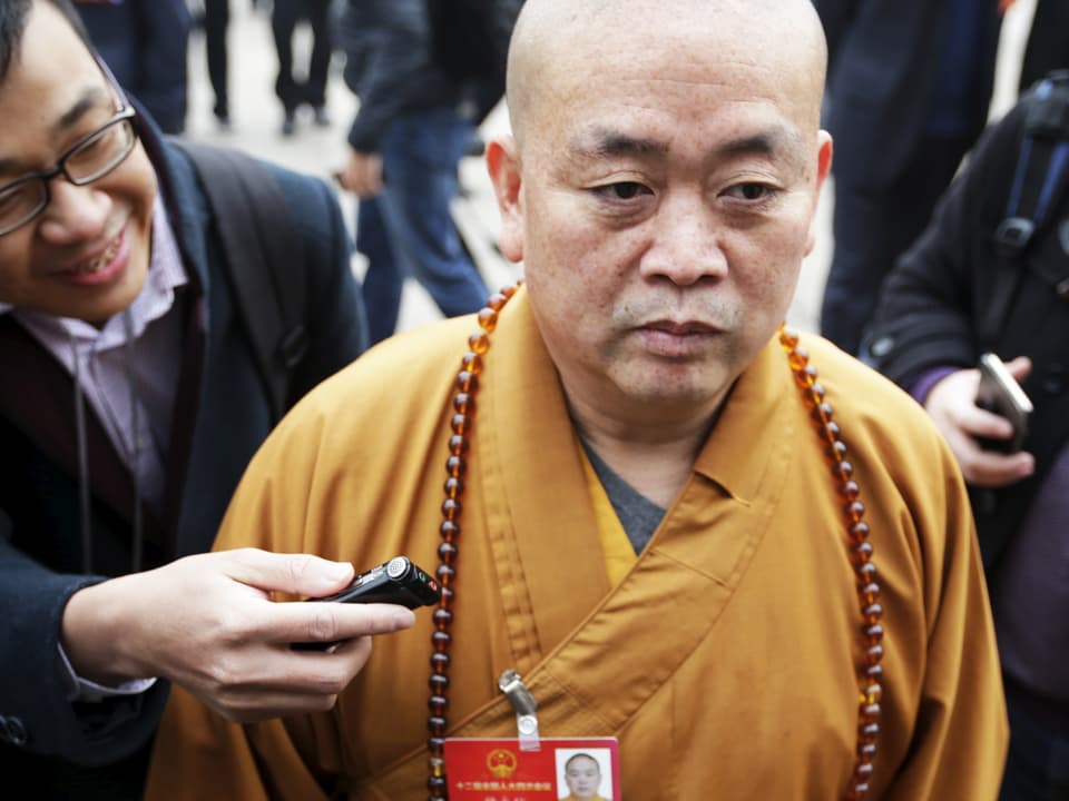 Shi Yongxin in Mönchskleidung, ein Mann hält ihm ein Aufnahmegerät hin.