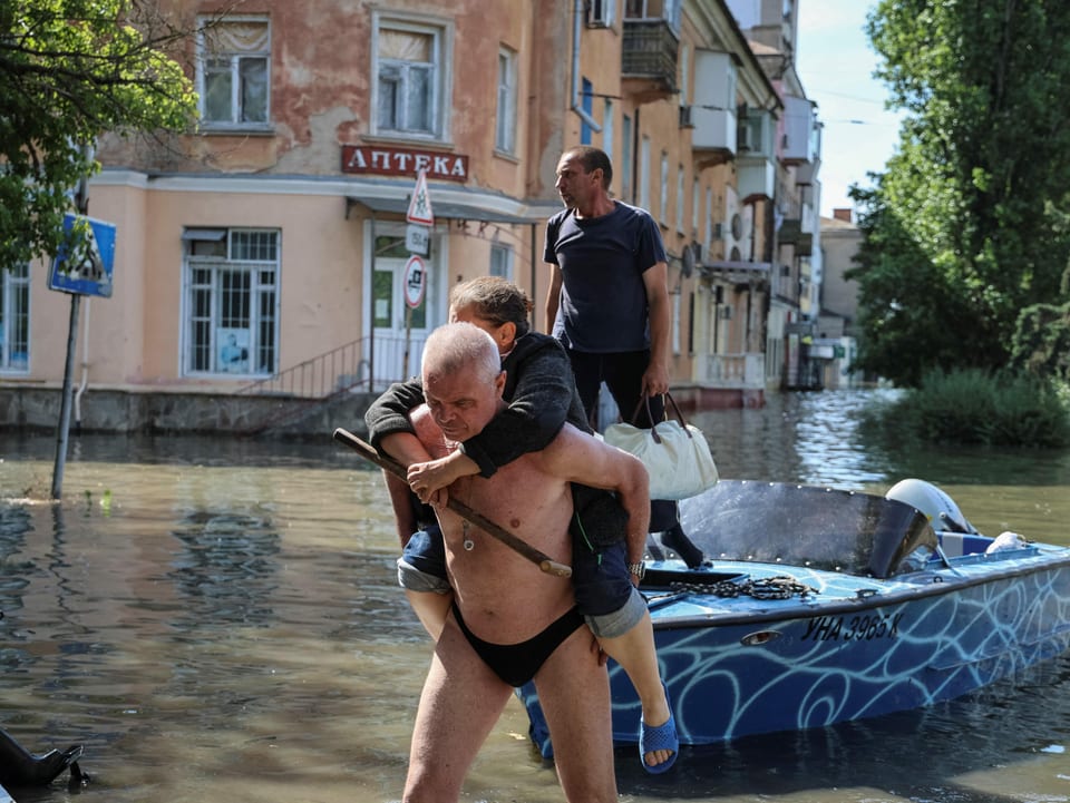 Blick auf eine überflutete Strasse. Ein Mann trägt eine Frau auf dem Rücken. Ein Boot liegt auf dem Wasser.