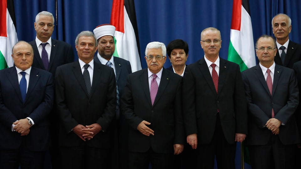 Mehrere Personen in Anzügen stehen vor palästinensischen Flaggen.