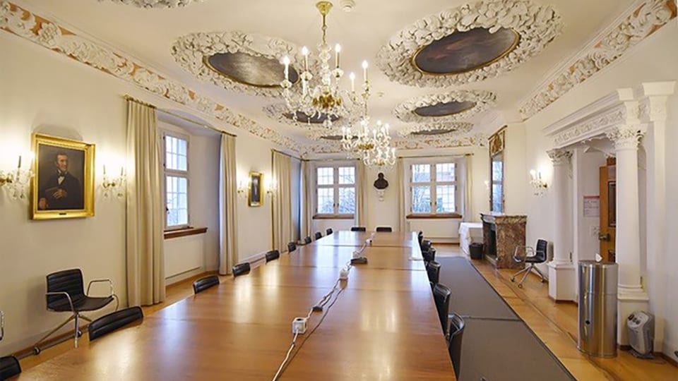 Eine Fotografie des Rektorats der Universität Zürich mit Stuck an der Decke, Kronleuchtern und einem langen Holztisch.