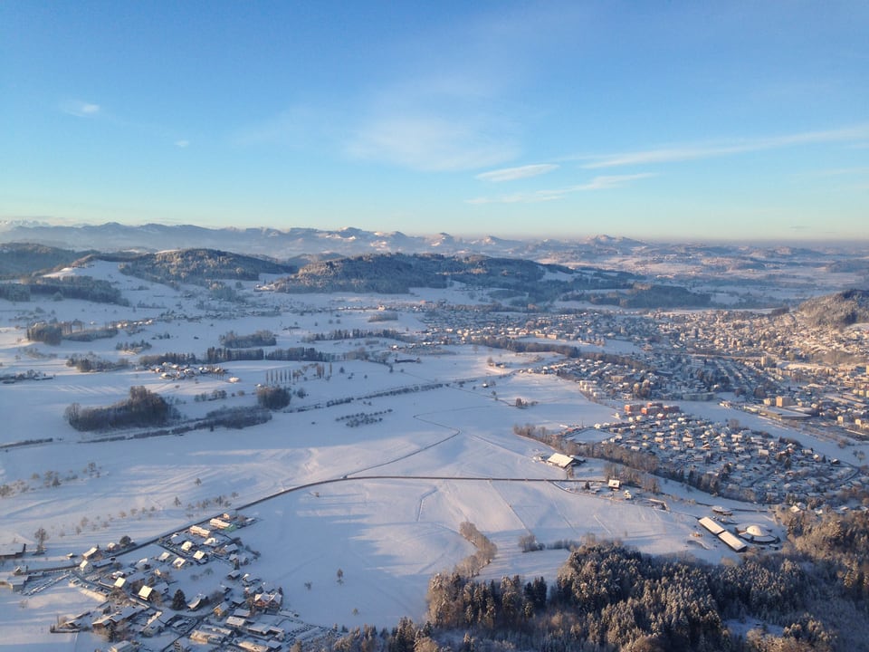 Luftbild auf die verschneite Landschaft des Mittellandes, dies bei Sonnenschein und blauem Himmel. Zu sehen sind grosse weisse Wiesen und kleine Dörfer. 