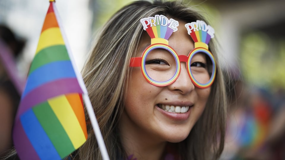 Ein Frau mit braunem langen Haar lächelt freundlich. Sie trägt eine Brille in Regenbogenfarben und schwenkt eine Fahne.