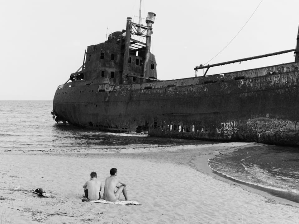 Zwei junge Männer sitzen am Strand vor einem Schiffswrack (Schwarz-Weiss-Bild)