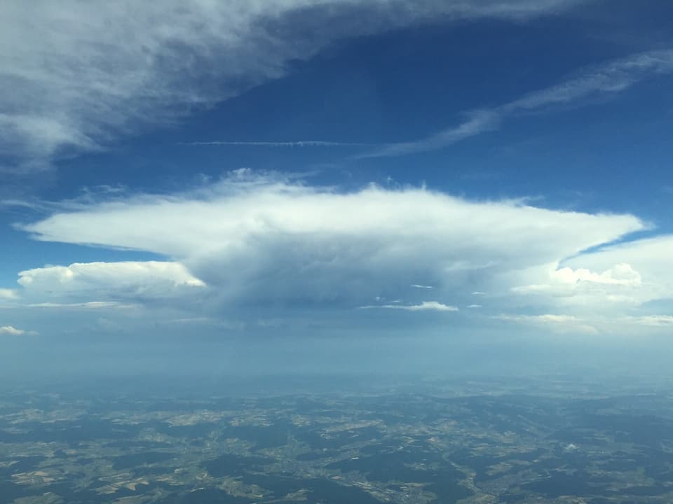 Luftaufnahme über das Schweizer Mittelland mit Muster aus Wald und Feldern auf eine grosse Gewitterwolken mit Ambossform. 