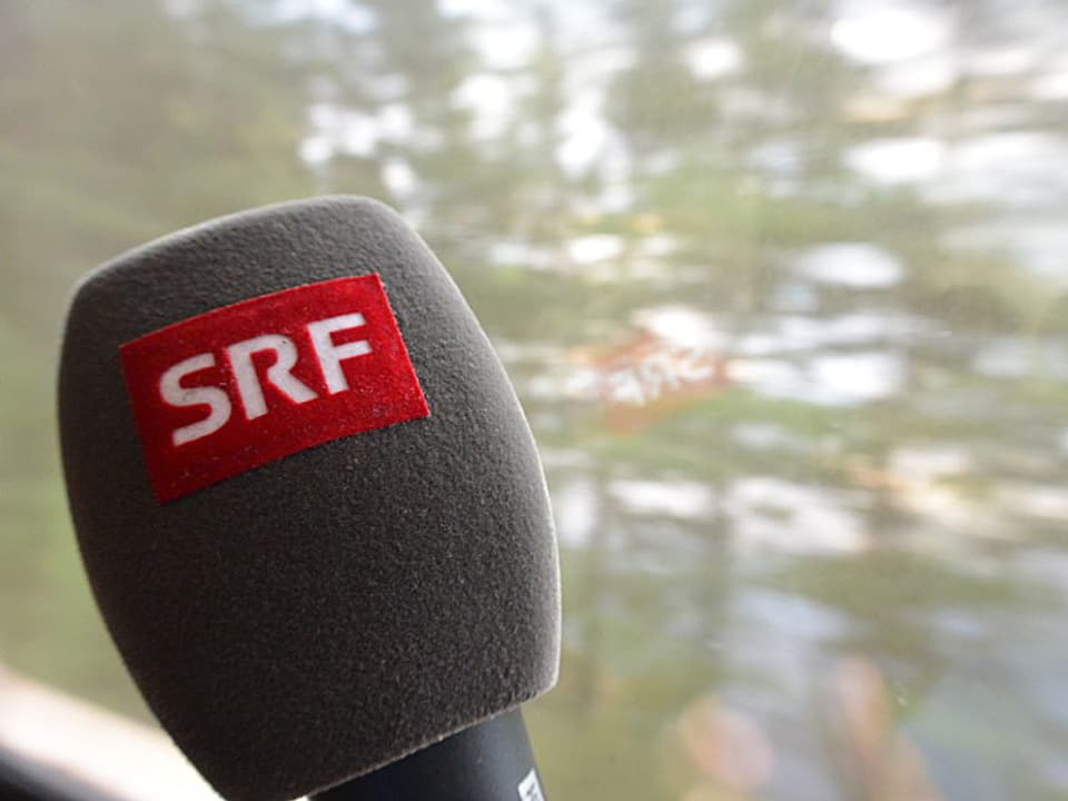 Ein SRF-Mikrofon vor dem Zugfenster, im Hintergrund verschwommene Bäume.