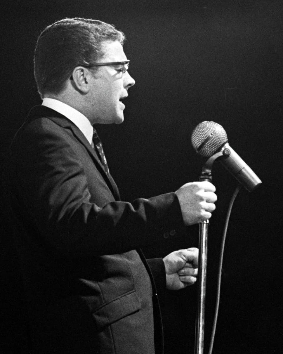 Schwarz-Weiss-Fotografie mit einem Mann im dunklen Anzug, der vor einem Mikrofon steht und singt.