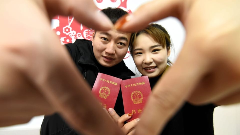 Junges chinesisches Paar hält Hände zu Herz zusammen, beide halten rotes Dokumentbüchlein