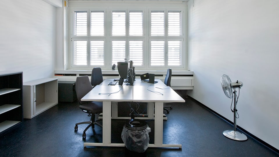 Ein ziemlich leeres Büro mit Ventilator, Tisch, Stühlen und Abfalleimer.