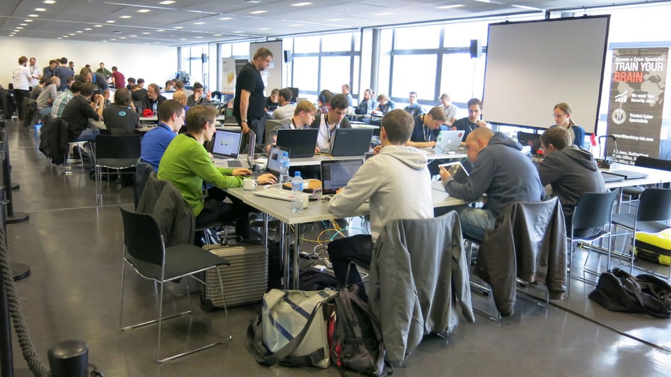 In einem Sitzungsraum sitzen viele junge Hacker an ihren Laptops.