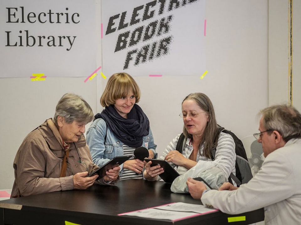 Zwei ältere Frauen und ein Mann sitzen an einem Tisch und lesen in einem E-Reader.