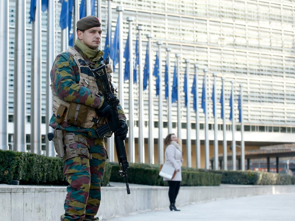Ein Soldat mit Gewehr vor der EU-Zentrale, im Hinergrund eine einsame Frau in Weiss