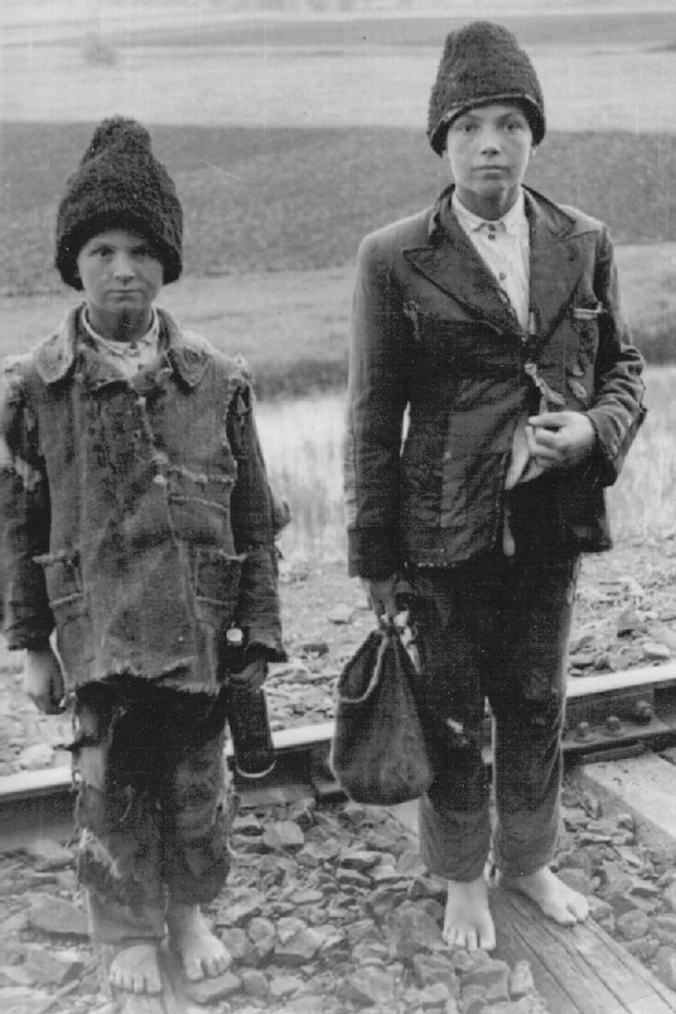 Zwei Kinder, barfuss und mit Mütze, zwischen Eisenbahngeleisen.