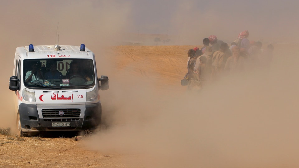 Eine Ambulanz fährt an einem Lastwagen voller Flüchtlinge vorbei, viel Sand wirbelt durch die Luft