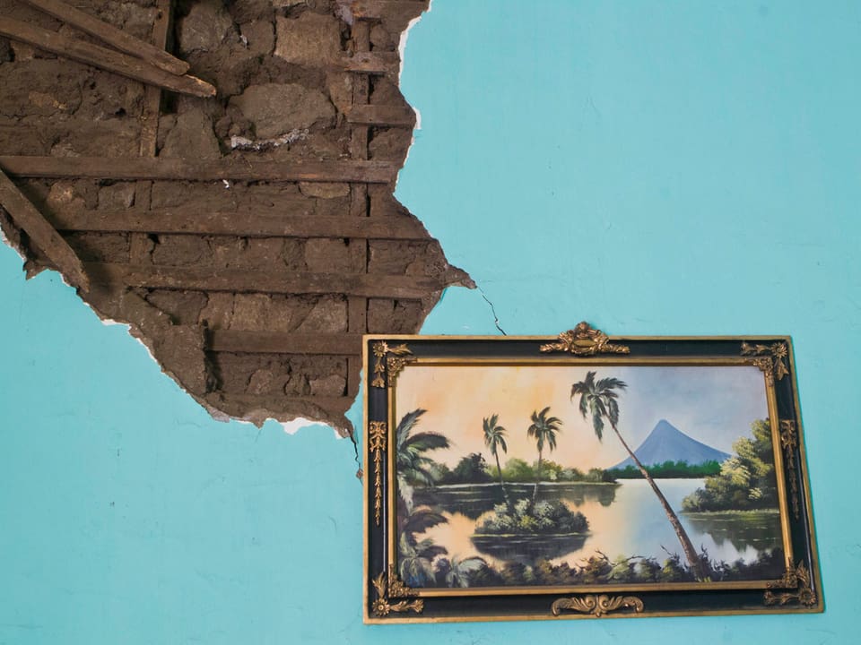 Ein Gemälde einer Landschaft mit Palmen, Inseln und einem Berg hängt an einer beschädigten, hellblauen Wand.