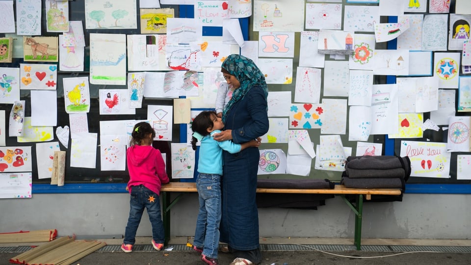 Frau mit Kopftuch und zwei kleine Kinder betrachten eine Wand voller Kinderzeichnungen.