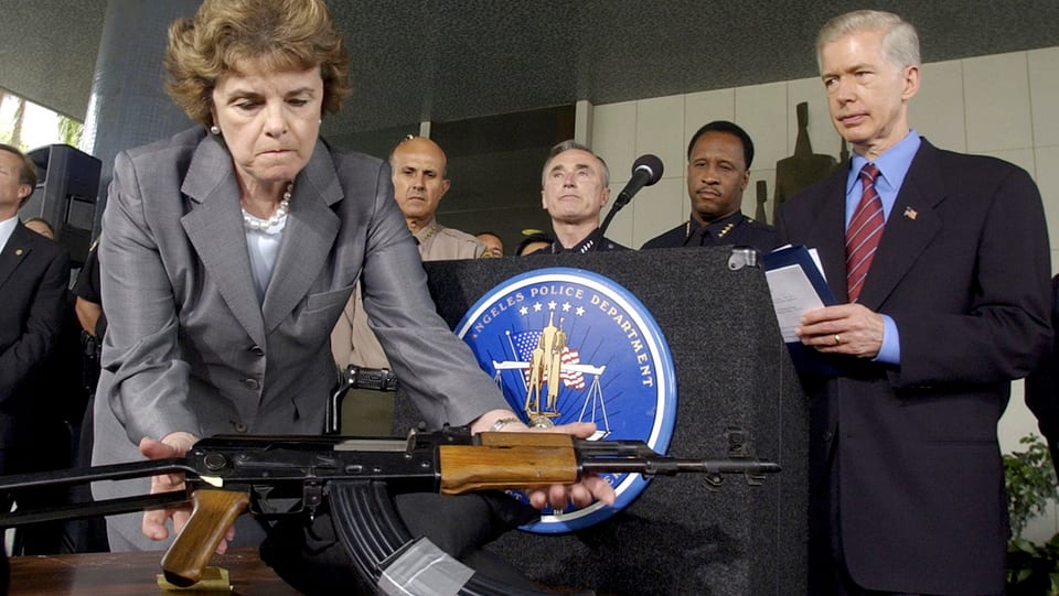 Dianne Feinstein, eine AK47-Sturmgewehr auf einen Tisch stellend. Im Hintergrund stehen Menschen.