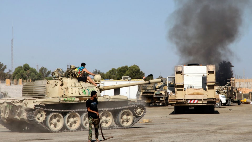 Ein Mann vor einem Panzer, im Hintergrund steigt Rauch auf.
