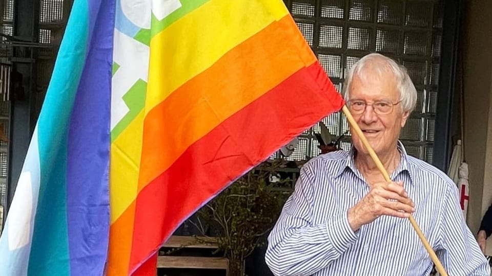 Älterer Mann trägt eine Regenbogen-Fahne in der Hand im Garten seines Hauses.
