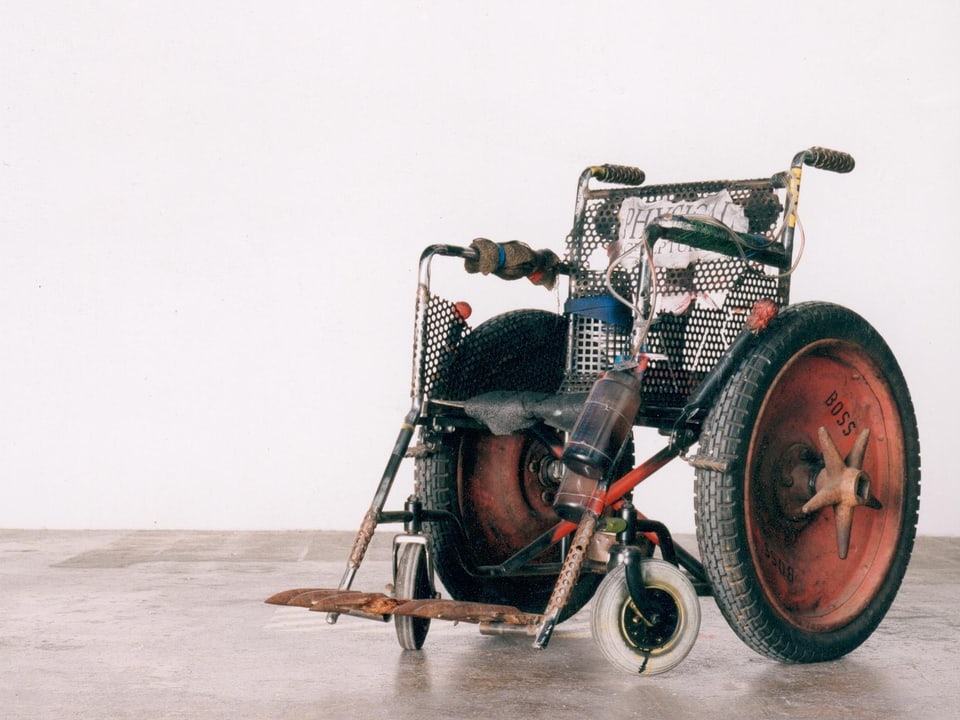 Metallener Rollstuhl mit Traktorenrädern, an dem unter anderem eine Flasche befestigt ist.