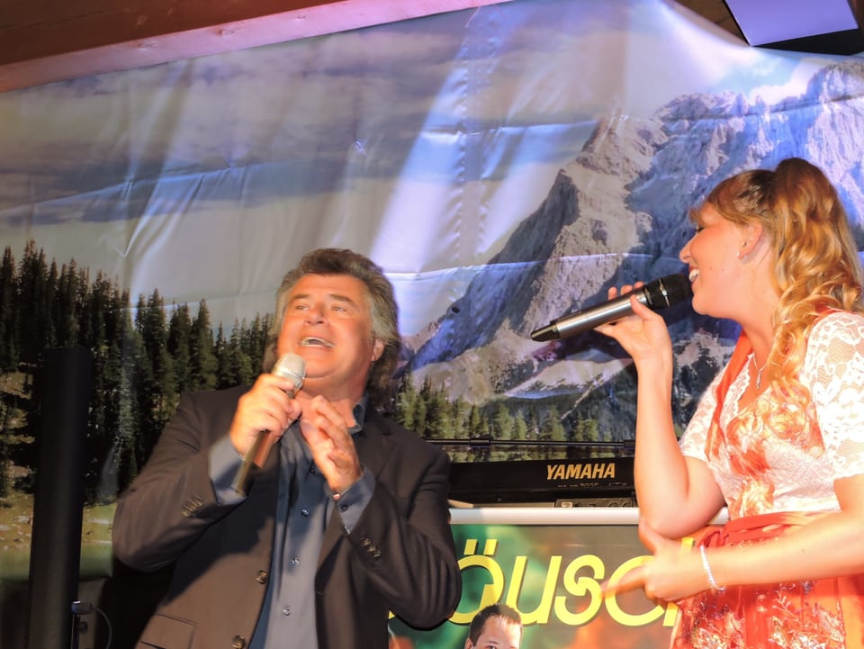 Zwei Sänger auf Bühne mit Poster von Berg als Hintergund.