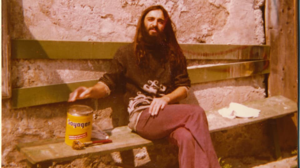 Ein Mann mit langen Haaren und Vollbart sitzt auf einer Bank. 