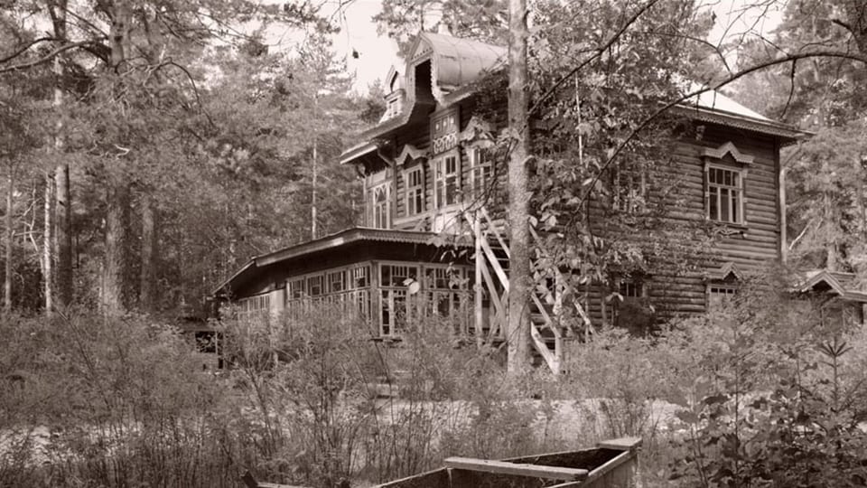 schwarzweiss Bild eines doppelstöckigen Holzhauses, davor Sträucher, dahinter ein Wald.