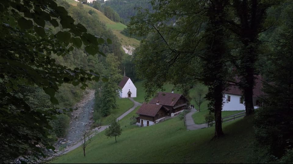 Landschaftsfoto unten Bauernhäuser und Kirche, rundherum grüne Hügel und Bäume.