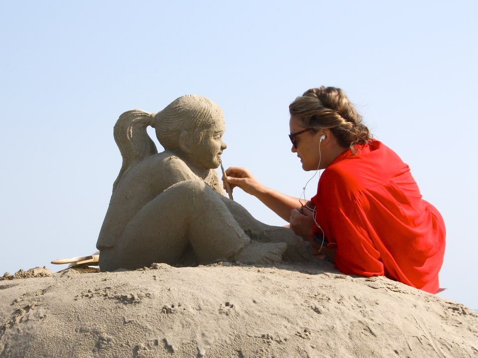 Eine Frau sitzt auf einem Haufen Sand und arbeitet an ihrer Sandskulptur. 