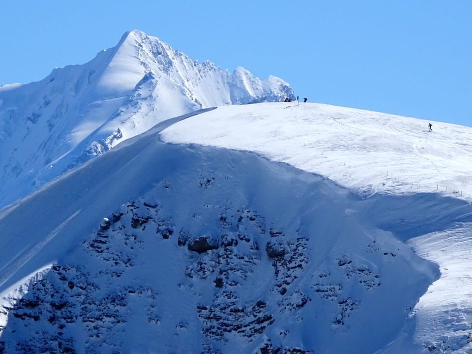Schneebedeckte Berge, darauf ein paar Tourenfahrer. Darüber blauer Himmel.