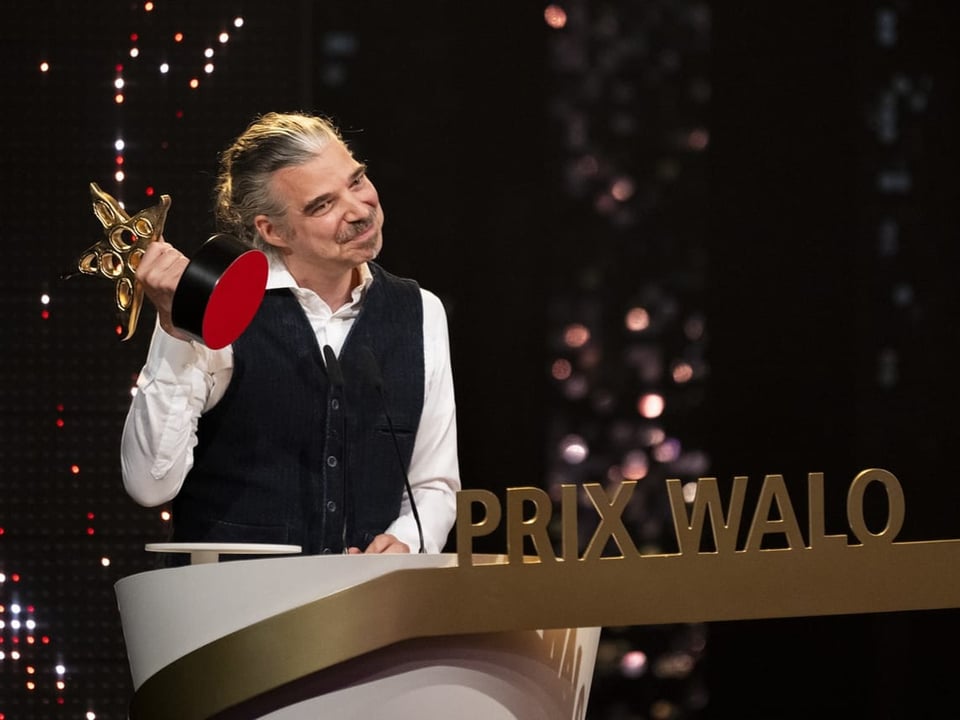 Mann mit Schnurrbart und Weste hält Preis bei der 'Prix Walo' Preisverleihung.
