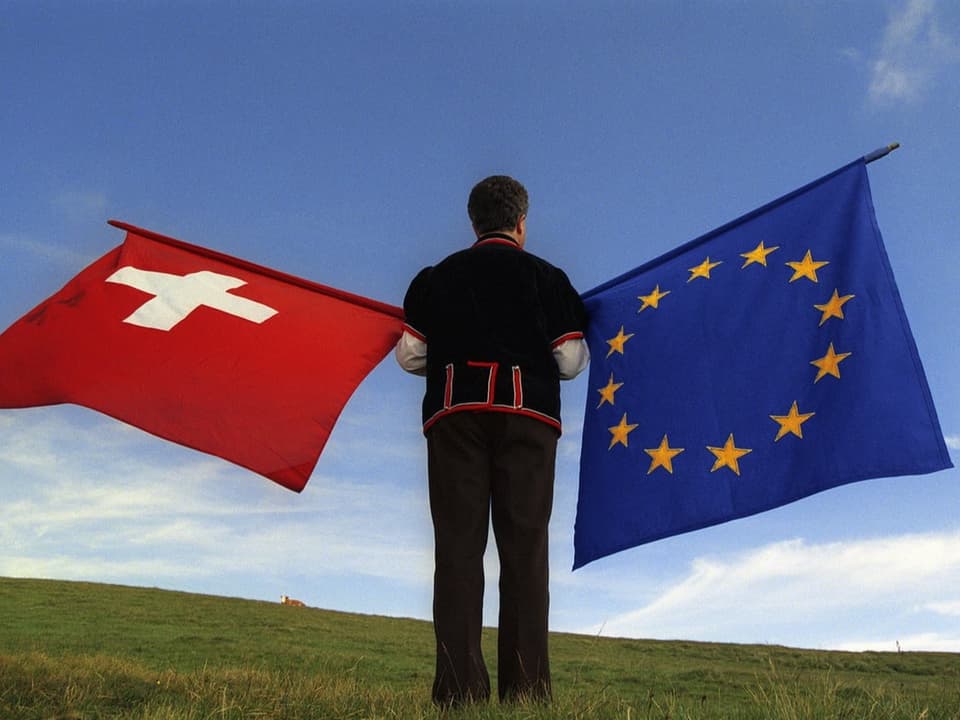 Ein Fahnenschwinger steht auf einer Wiese und hält je eine Schweizer Fahne und eine EU-Fahne.