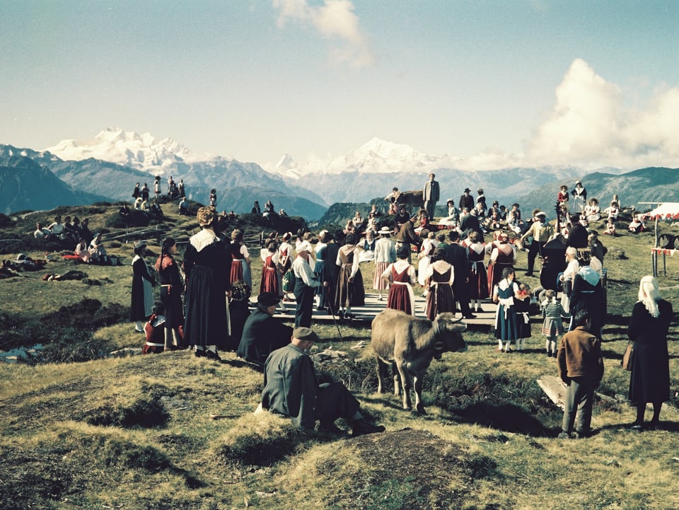 Filmset auf einer Alpwiese vor Alpenpanorama. Viele Statisten stehen auf einer Wiese. Zwischen ihnen zwei Kühe.