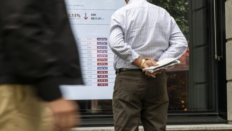 Mann steht vor einem Bildschirm, der die Börsenkurse abbildet.