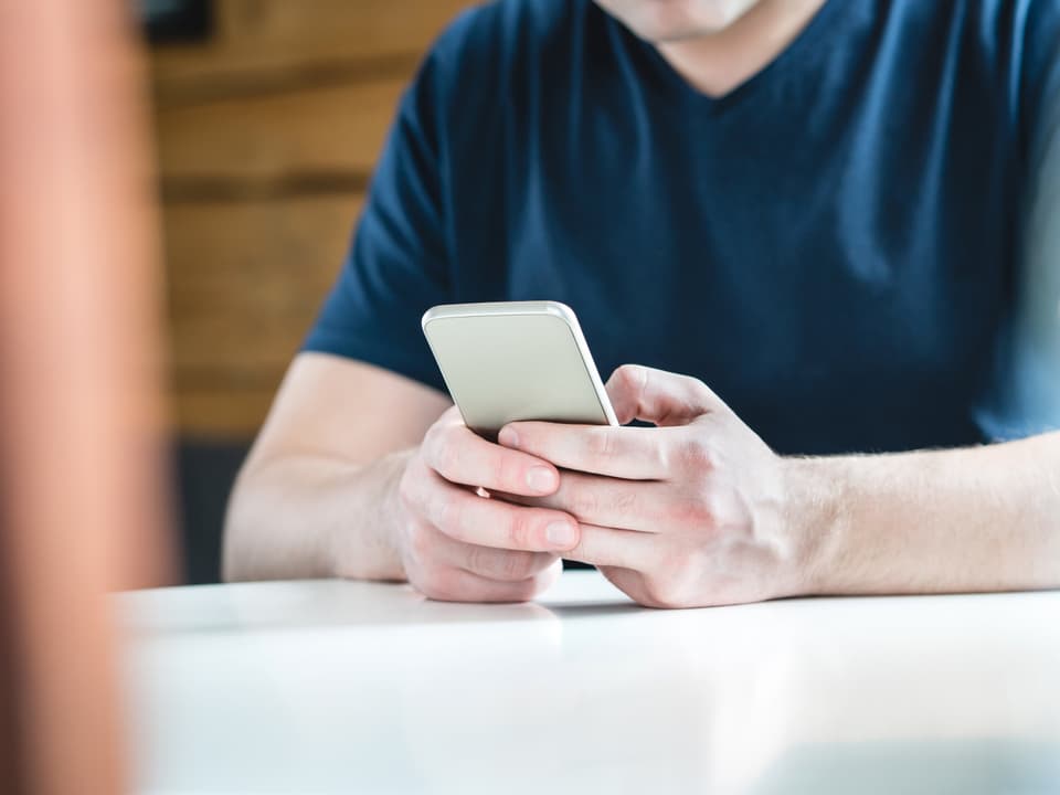 Ein Mann sitzt mit einem Smartphone an einem Tisch und tippt auf den Bildschirm.