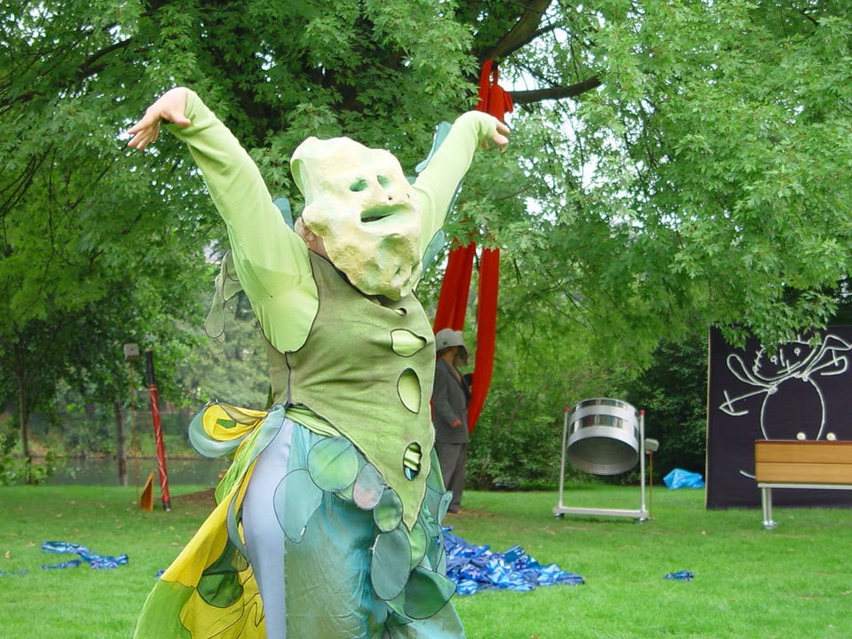 Eine verkleidete Figur mit Maske, auf einer Wiese tanzend.