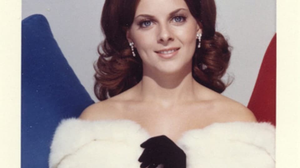 Eine weisse Frau mit braunen Haaren und blauen Augen trägt einen weissen Pelz Oberteil und schwarze Handschuhe.