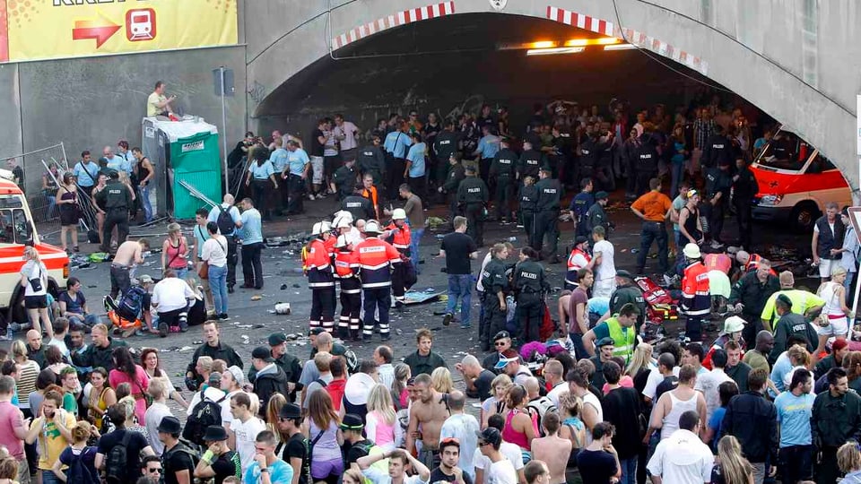 Blick in den Zugangsbereich der Loveparade mit dem Tunnel, in dem die Massenpanik stattfand: Viele Menschen stehen herum, in der Mitte Rettungssanitäter in roten Jacken.