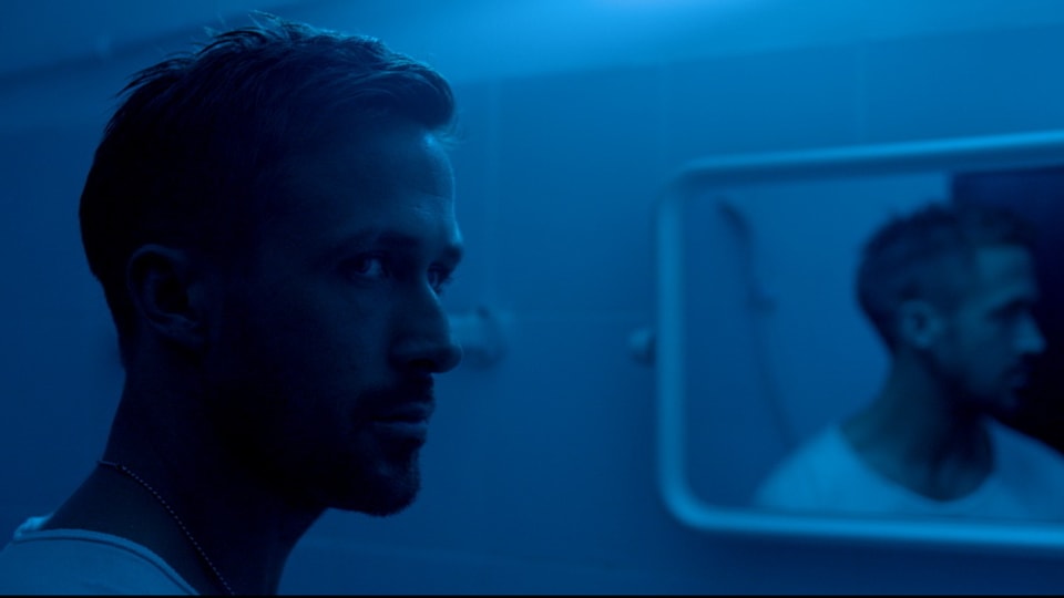 Hauptdarsteller Ryan Gosling in, blaues Neonlicht getaucht, steht vor einem Spiegel.