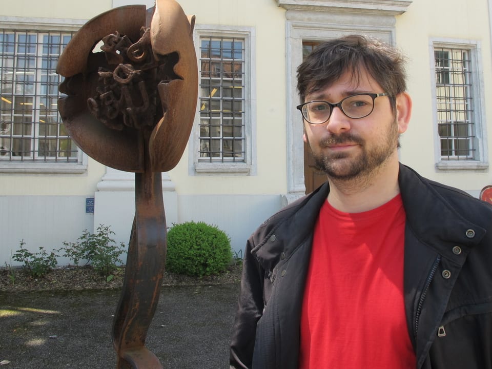 Bild von Patrick Bohrer, wie er vor dem Eingang der Bibliothek steht. Er trägt eine schwarze Jacke mit einem roten T-Shirt darunter, und hat eine Brille und einen 3-Tage-Bart. Links von ihm steht eine Eisenskultpur.