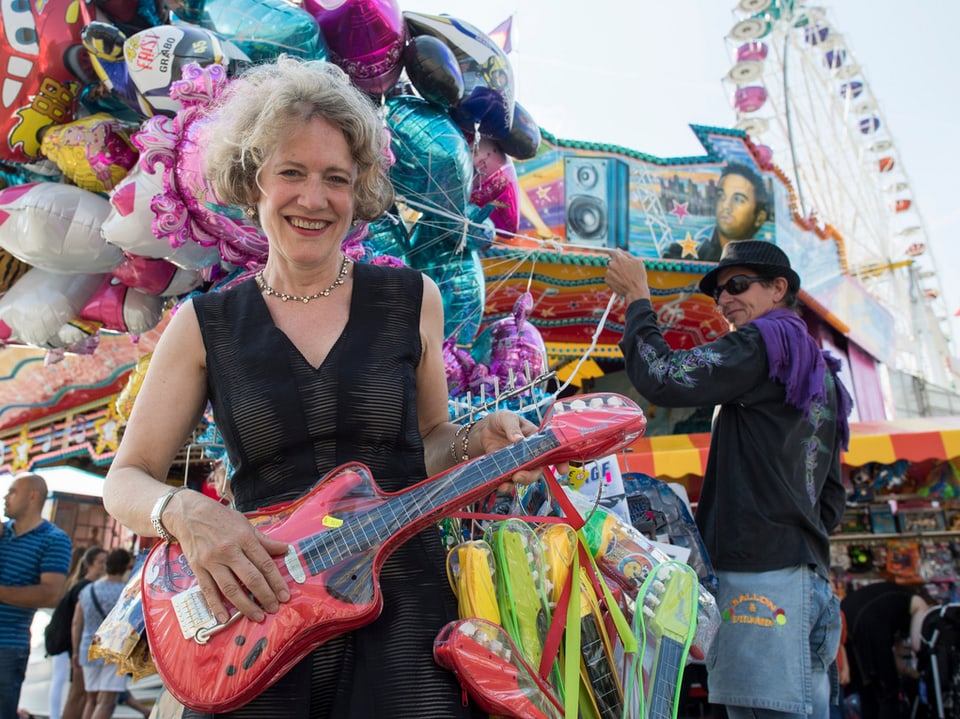 Eine Frau in Kleid hält eine Plastik-Gitarre in den Händen und lächelt in die Kamera. Im Hintergrund ein Riesenrad.