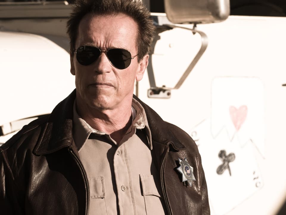Arnold Schwarzenegger in einer Polizei-Lederjacke und dunkler Sonnenbrille. 